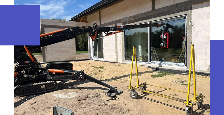 Długa, czarna rama okienna lub drzwiowa jest podnoszona przez maszynę z wysięgnikiem i przyssawkami, obok niedokończonego białego budynku mieszkalnego.